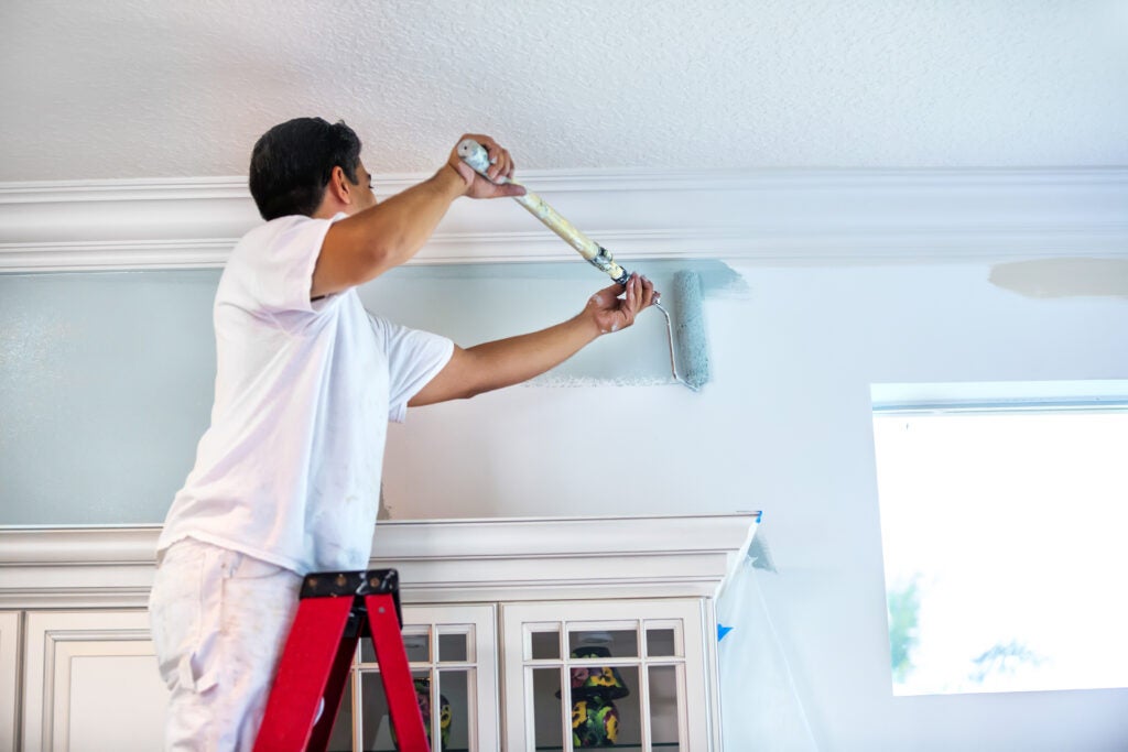 A housepainter stands on a ladder to paint the walls inside a residential home. 