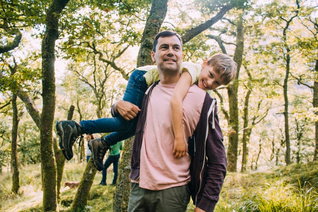 Little boy is smiling at the camera as he is being fireman carried by his father through the woodlands.